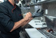 Chef de jaqueta preta calçando luvas em uma cozinha profissional de inox