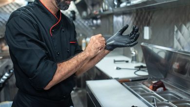 Chef de jaqueta preta calçando luvas em uma cozinha profissional de inox