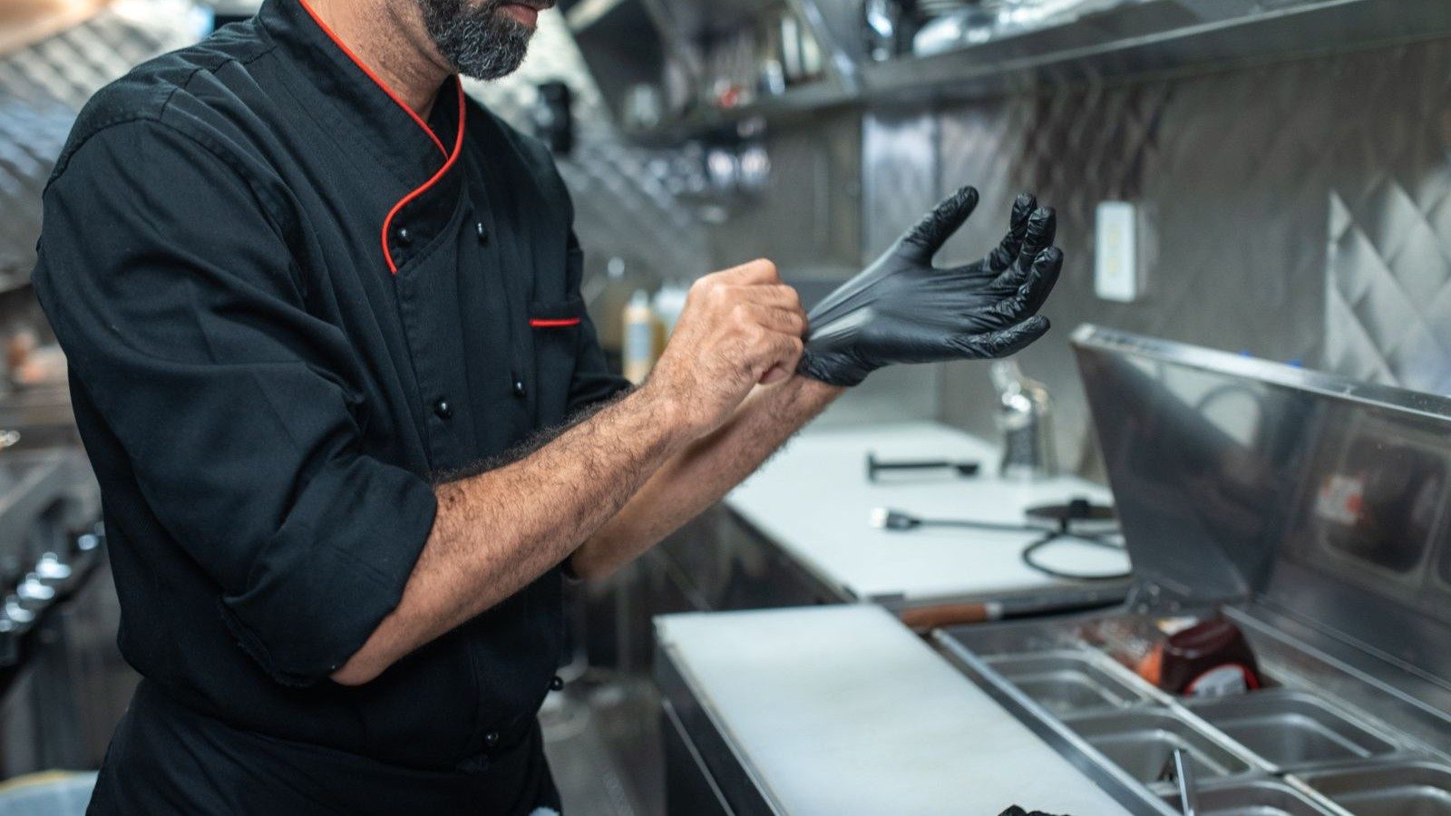 Chef de jaqueta preta calçando luvas em uma cozinha profissional de inox
